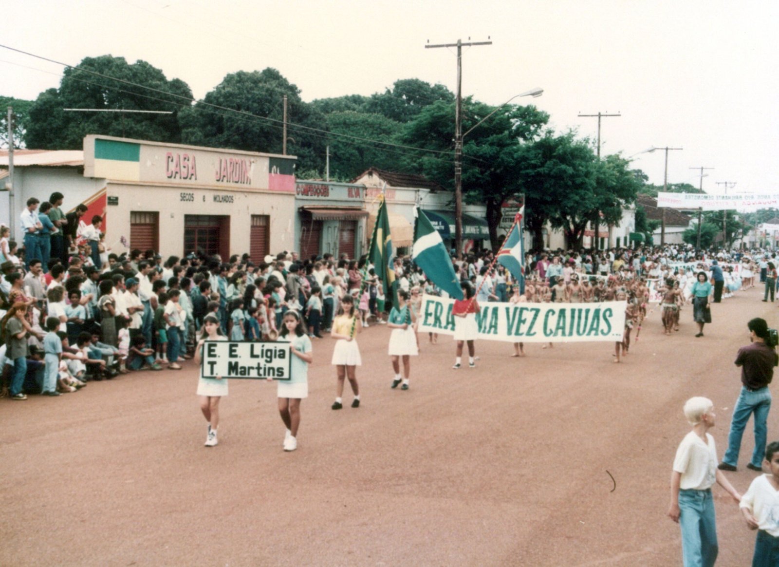 Desfile cívico na cidade de Rio Brilhante com pessoas caminhando em estrada de terra.